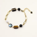 Gold chain bracelet with tiger's eye, obsidian, pearl, and iridescent abalone stones on cream background.
