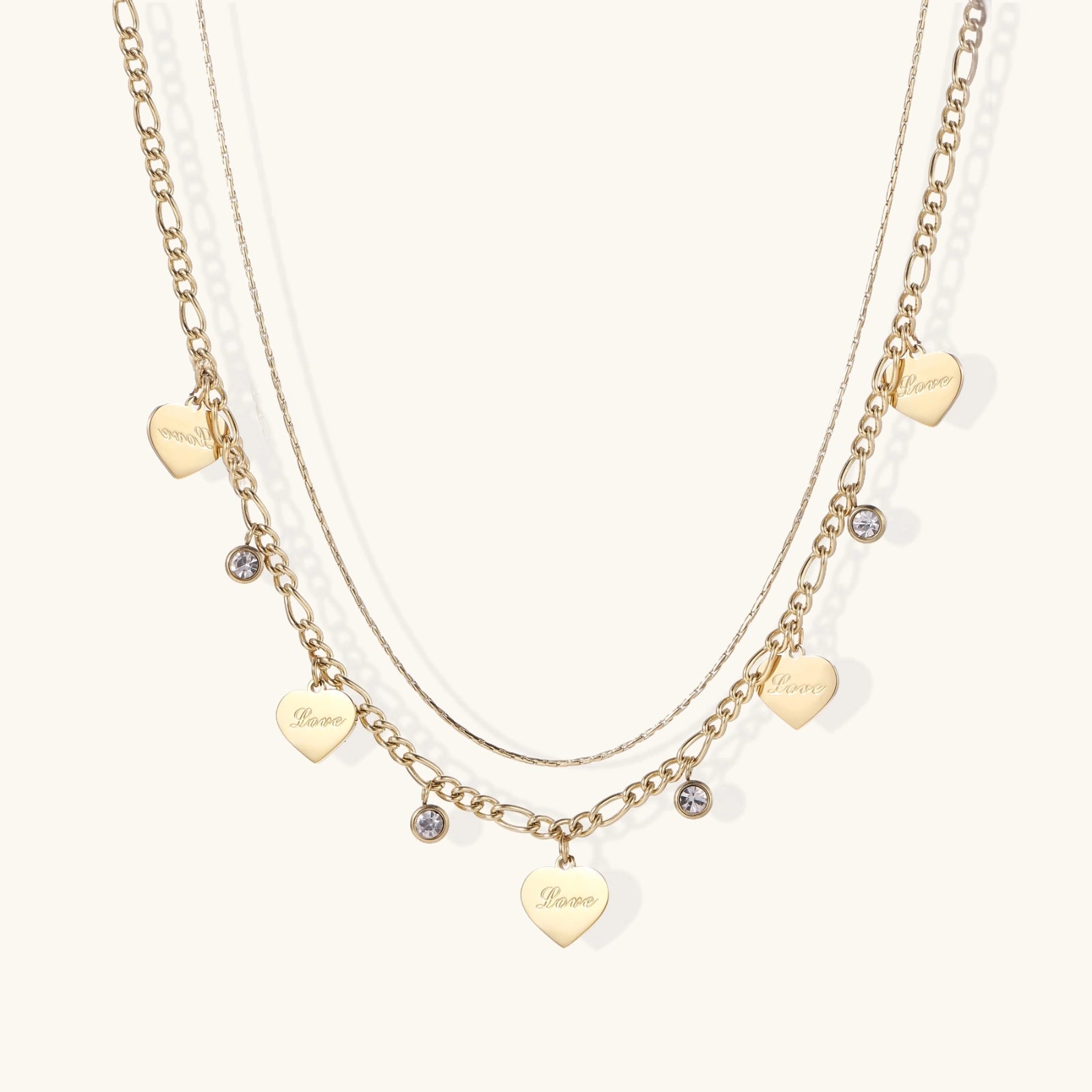 Gold layered necklace set with engraved heart pendants and crystal accents on cream background