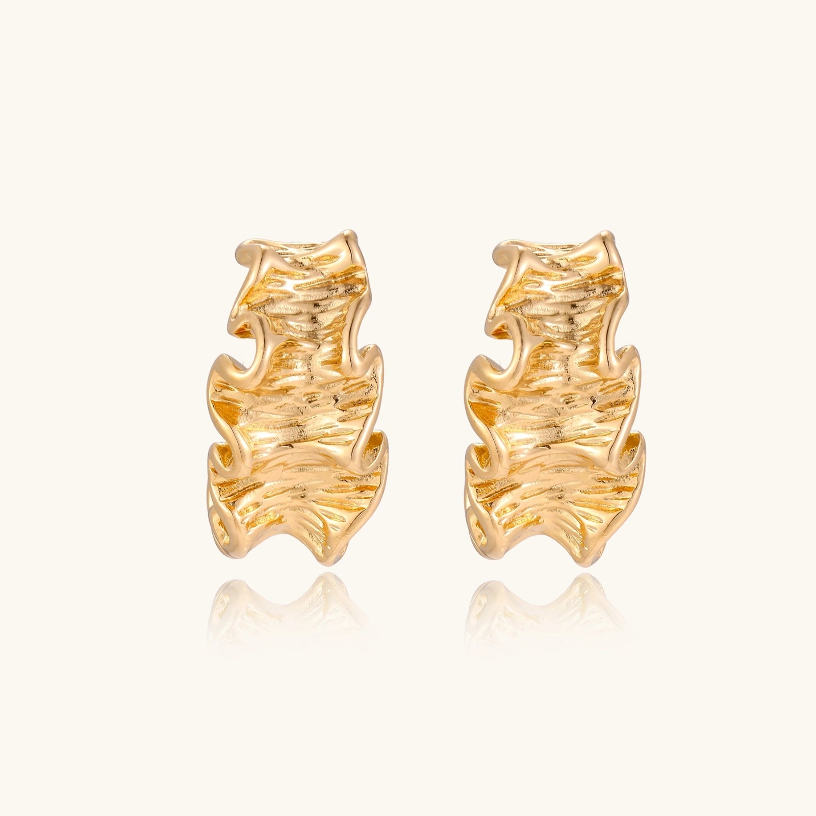 Pair of gold huggie hoops with sculpted wave texture and dimensional ripples on neutral background.