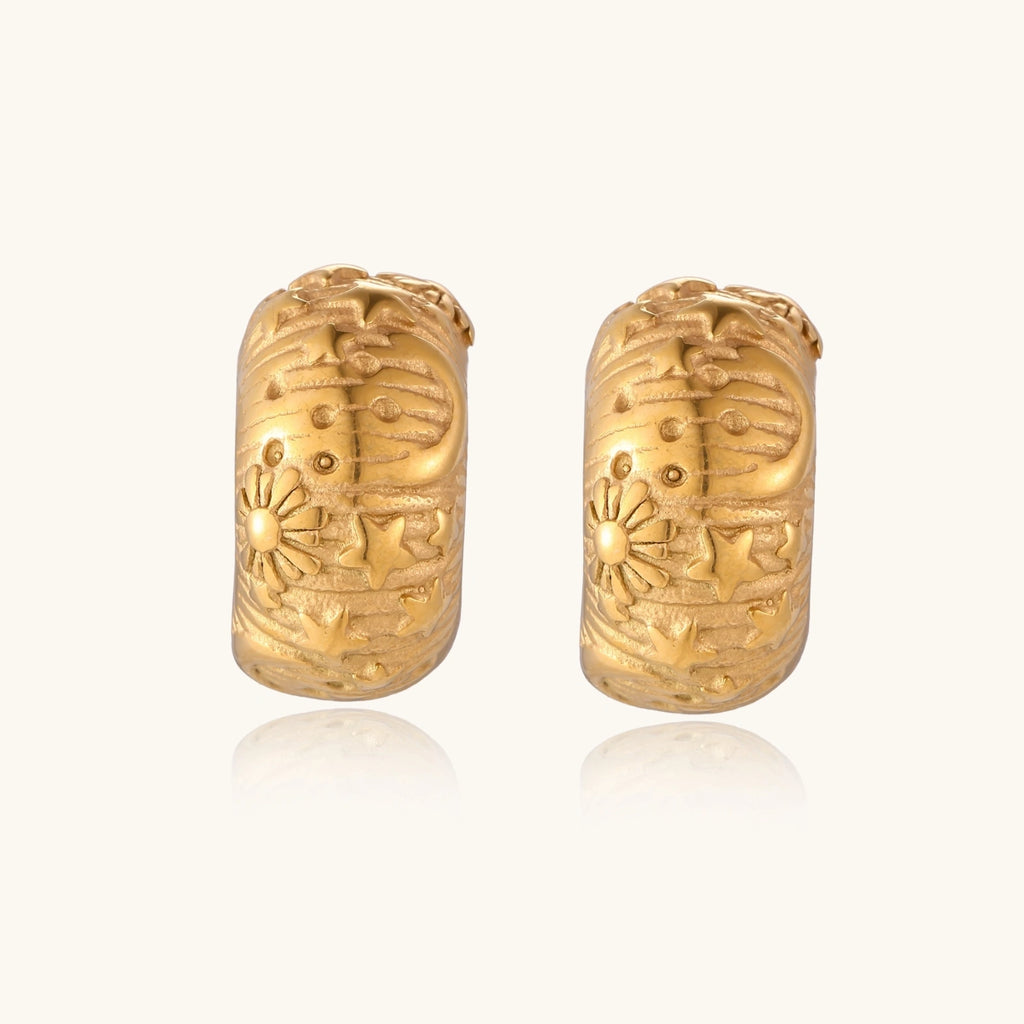 Wide gold hoop earrings with embossed stars and sun motifs on cream background, showing dimensional relief details.